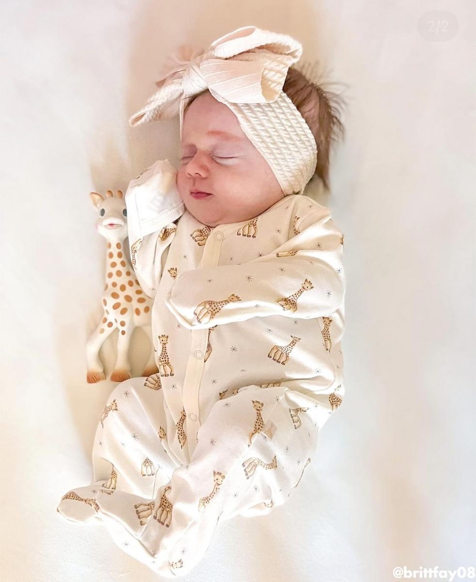 Baby wearing Kissy Kissy giraffe print footie pajamas and a large bow headband next to a giraffe toy