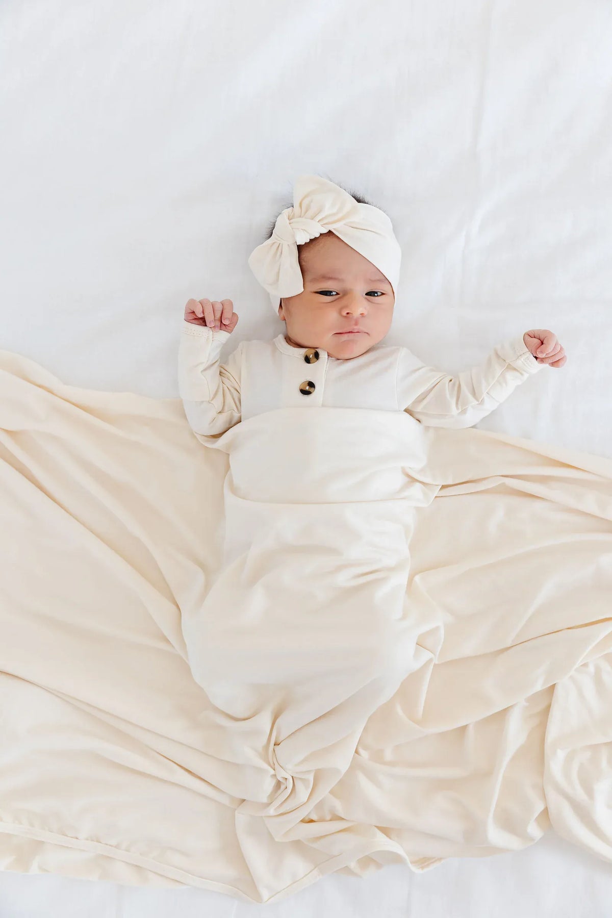 Newborn wearing Lou Lou and Company cream knotted headband and button front swaddle gown on white bedding