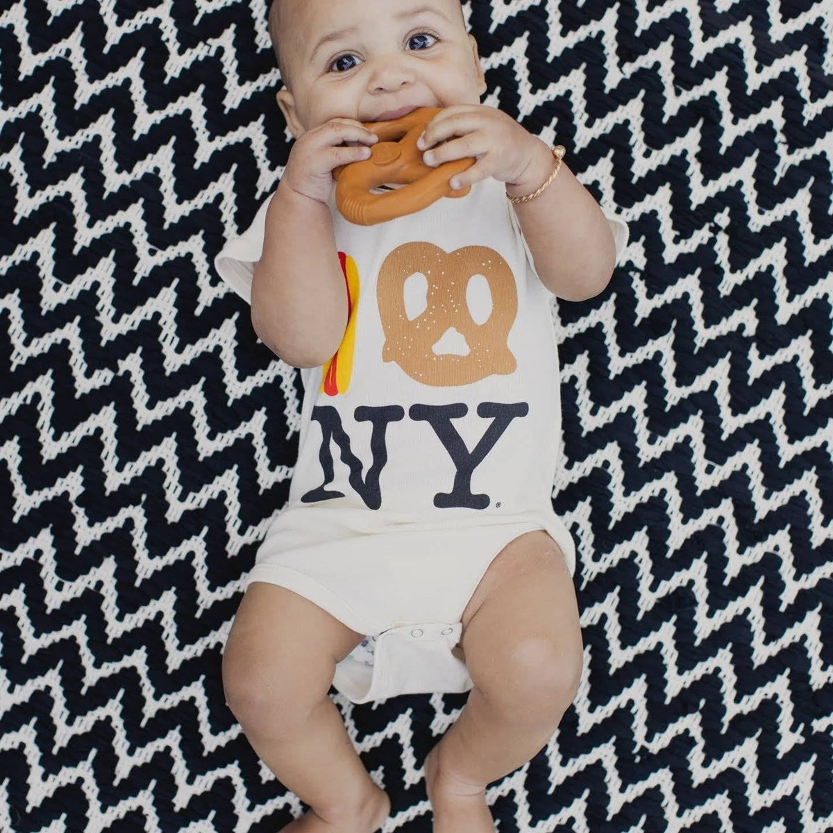 Baby wearing a cream colored onesie with an I love NY graphic while chewing on a pretzel shaped teether