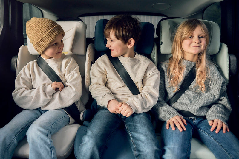 Three children smile and chat while securely seated in Axkid One 3 and Up car seats during a cozy car ride.