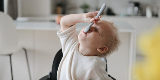 A baby gleefully tries to eat using a gray spoon, capturing the joy and messiness of learning to self-feed.
