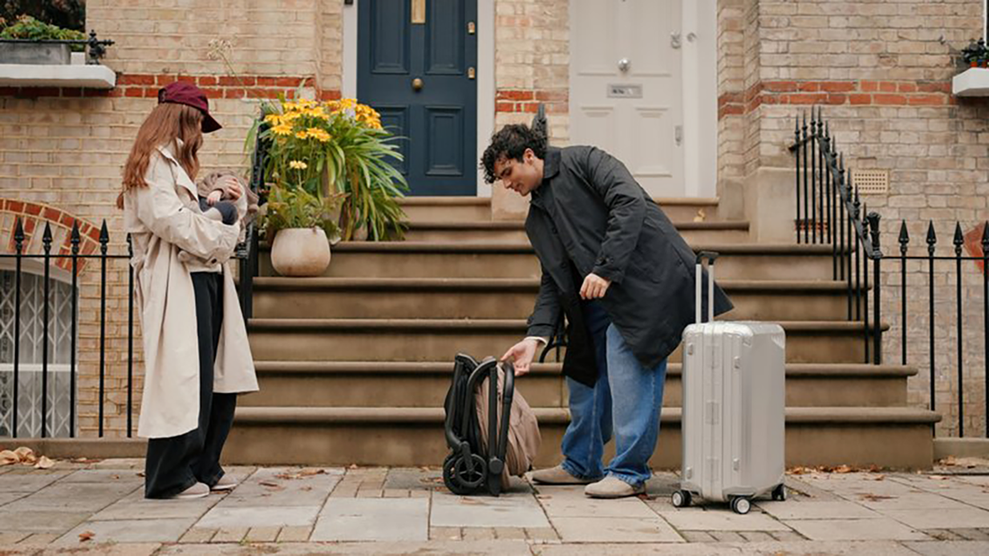 A young family prepares for travel outside their brick home. The father folds a Silver Cross Nia stroller next to his luggage, while the mother holds their baby nearby.
