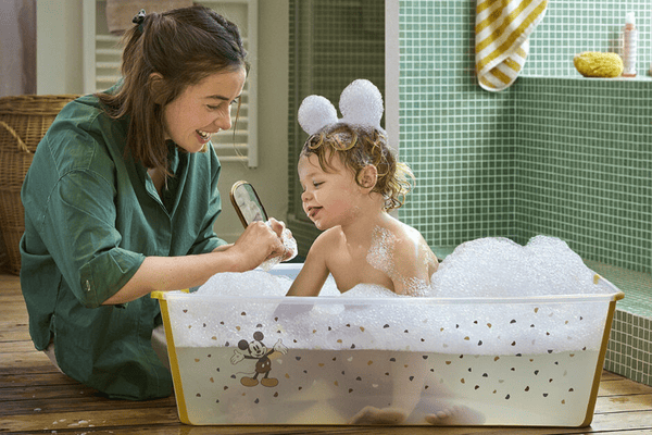 A smiling mother bathes her toddler, who has bubbles on their head, in a Mickey Mouse tub, creating a playful and relaxed atmosphere.

