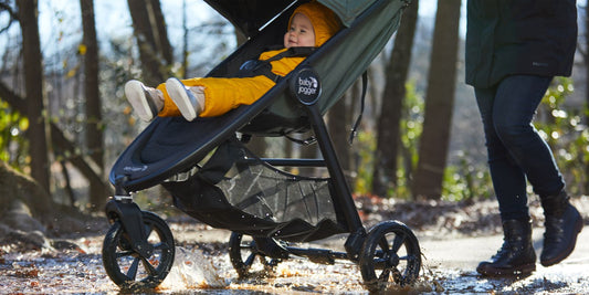 A happy baby in a Baby Jogger stroller splashes through puddles on a wooded path with its parent, enjoying an adventurous and affordable ride.
