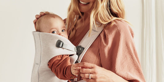 A mother smiles while holding her baby in a light grey carrier, creating a sense of love and security.
