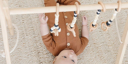 A baby in a brown ribbed onesie lies under a wooden play gym, reaching for toys, conveying a peaceful and playful mood.
