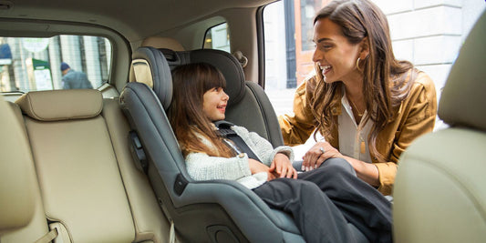 A mother smiles at her daughter, who is safely buckled in a modern convertible car seat in the back of a car.
