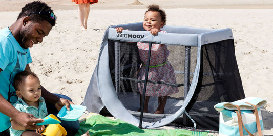 A happy family enjoys a sunny day at the beach, with a baby safely playing in a breathable Aeromoov travel cot.
