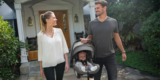 A young family smiles while strolling away from their house with their baby in an infant car seat.

