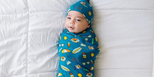 A baby swaddled in a blue summer-themed blanket and matching hat rests peacefully on a white quilt, calm and content.
