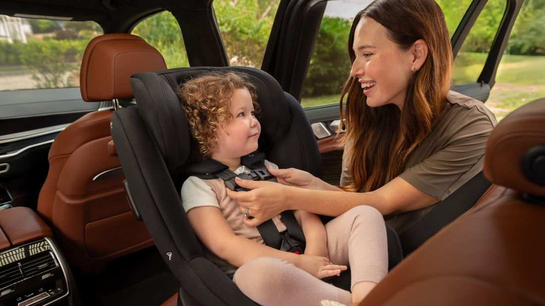 A mother smiles as she secures her young daughter in a black Nuna RAVA car seat inside a luxurious vehicle.
