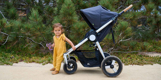 A sweet toddler in a yellow romper stands with a stroller, holding flowers, on a nature walk, highlighting eco-friendly family fun. 
