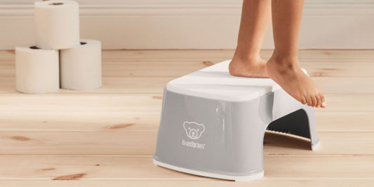 A child stands on a gray BABYBJÖRN step stool in a bathroom, reaching for the toilet paper beside them.
