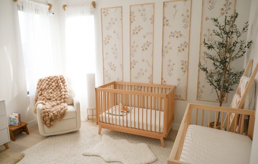 A serene nursery with two wooden cribs, floral wall art, soft lighting, and a cozy armchair invites a sense of calm and cleanliness.
