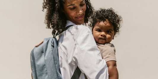 A smiling mother with curly hair holds her baby while wearing a stylish backpack, suggesting practical and fashionable diaper bag options.
