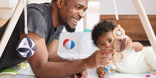 A smiling dad encourages his baby during tummy time, pointing to a wooden toy suspended from a play gym.
