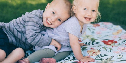Two happy young children cuddle on a floral blanket in a grassy outdoor setting, radiating warmth and sibling affection.