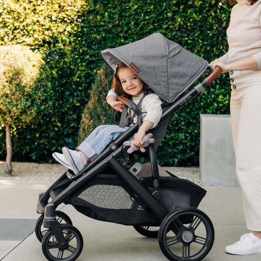 A happy toddler sits in the UPPAbaby Vista stroller, ready for a walk. Lush greenery forms the backdrop.
