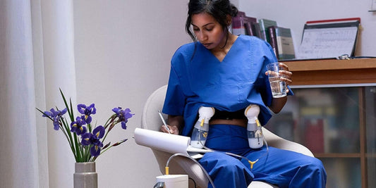 A woman in blue scrubs sits using a breast pump, multitasking with paperwork and water, capturing the reality of pumping on the go.
