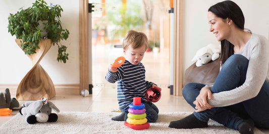 A tender scene of a toddler happily playing with a ring stacker toy as his mother lovingly watches in their baby-proofed living room.
