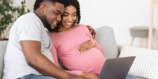 A smiling pregnant couple sits on the couch, looking at a laptop while creating their baby registry.

