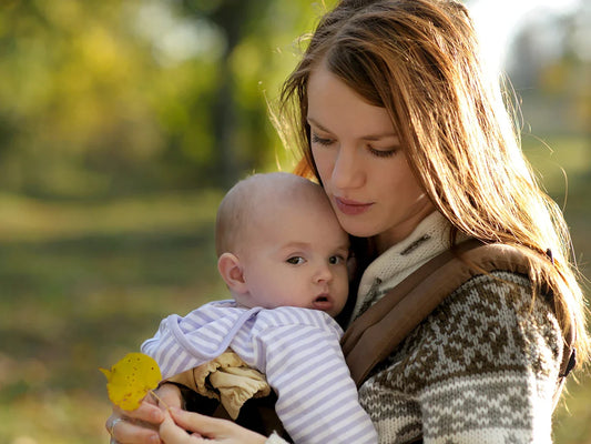 A mother lovingly gazes at her baby, held close in a carrier amidst an autumn park, portraying the tender bond enhanced by babywearing.
