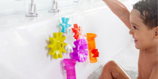 A toddler smiles in the bath, happily pouring water onto colorful, interconnected gear bath toys attached to the tub.
