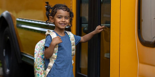 A smiling girl with a backpack touches a school bus door, conveying the excitement of going back to school.

