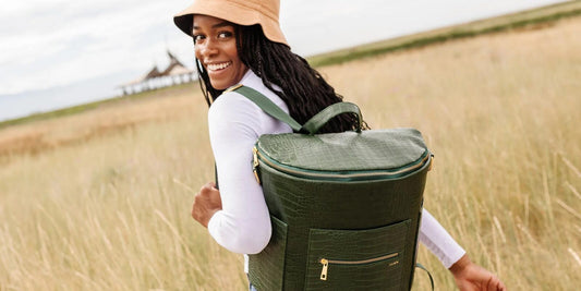 A smiling woman walks through a field with a stylish green backpack-style diaper bag, evoking a sense of freedom and preparedness.
