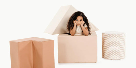 A girl with curly hair peeks out of a foam block house, conveying playful summer fun with indoor toys.