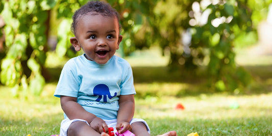 A happy baby sits on the grass in a blue shirt with an octopus, enjoying a bright spring day.
