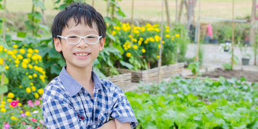 A smiling Asian boy in a garden, wearing glasses and a plaid shirt with his arms crossed.
