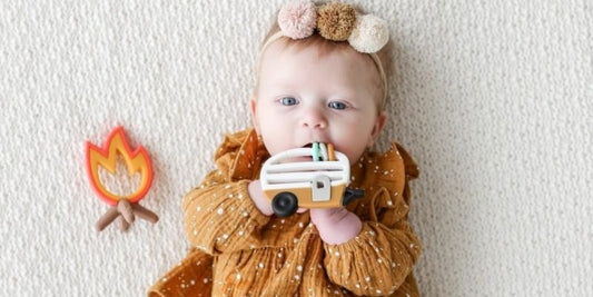 A cute baby, wearing a pompom headband and mustard-colored dress, chews on a camper teether while lying on a cozy blanket.