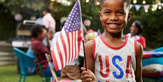 A smiling young boy proudly waves an American flag at a festive backyard July 4th gathering.
