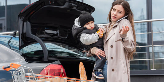 A stressed mother on her phone holds a baby while unloading groceries from the car, capturing the chaos of running errands. 
