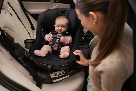 Smiling baby in black Nuna REVV maxx car seat, mother adjusting harness in car, cozy and secure.