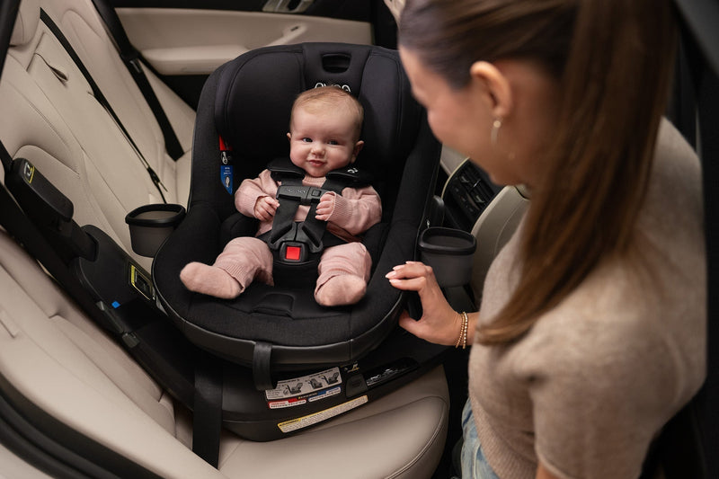 Smiling baby in black Nuna REVV maxx car seat, mother adjusting harness in car, cozy and secure.