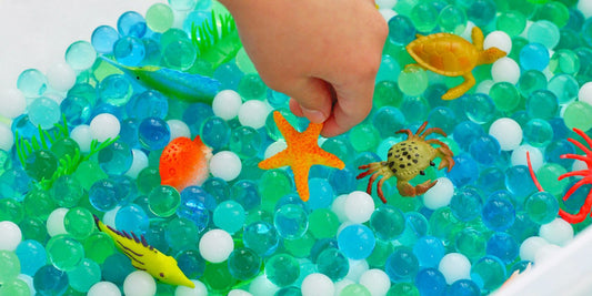 A child's hand picks up an orange starfish in a sensory bin filled with ocean-themed toys and water beads.