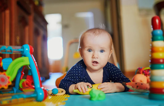 A curious baby with bright blue eyes lies on a colorful playmat surrounded by toys, engaged in tummy time.