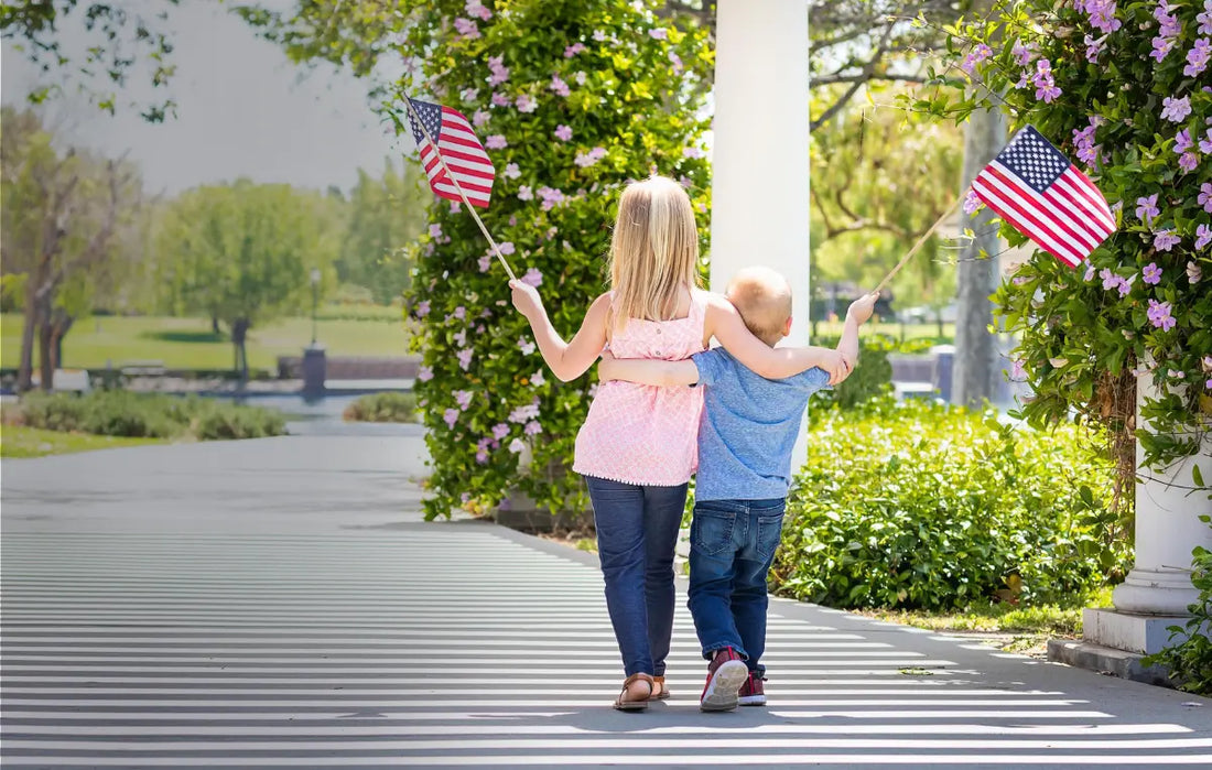 Two young children walk arm in arm beneath a flower-covered arbor, each holding a small American flag, on a sunny day outdoors.