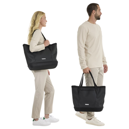 Black Thule Chasm Gear Tote 30L by Thule, shown in lifestyle side view with models carrying, featuring durable fabric, front logo, and dual handles.