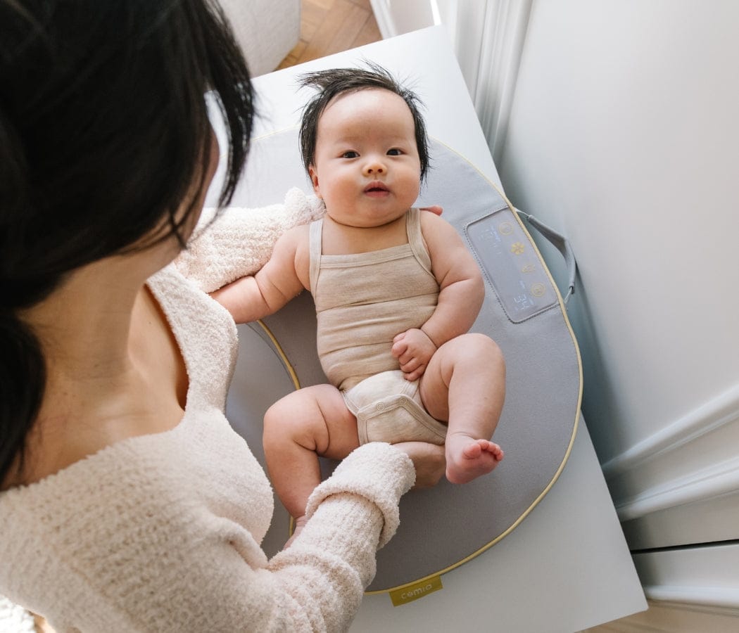 A baby lies on a gray changing pad with yellow trim, looking at a woman wearing a cream-colored sweater. "comia" is visible on the pad.
