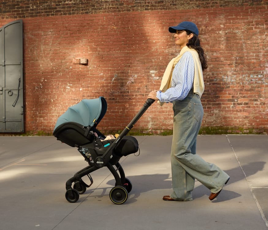 Woman in blue cap and jeans pushes black stroller with teal canopy along sidewalk beside red brick wall.