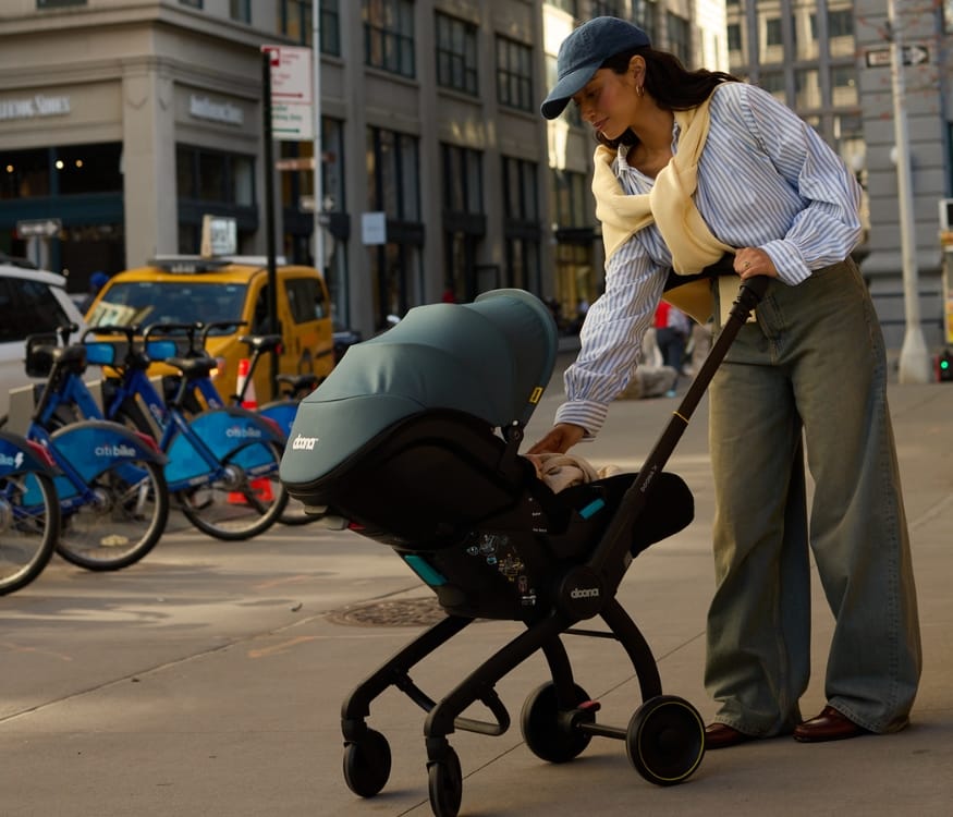 Woman in blue cap and striped shirt adjusts baby in teal stroller on city sidewalk, with Citi Bikes and yellow taxi in background.