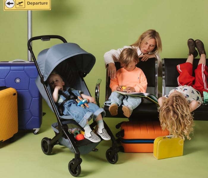Child sitting in a blue stroller next to a woman and children in an airport terminal setting