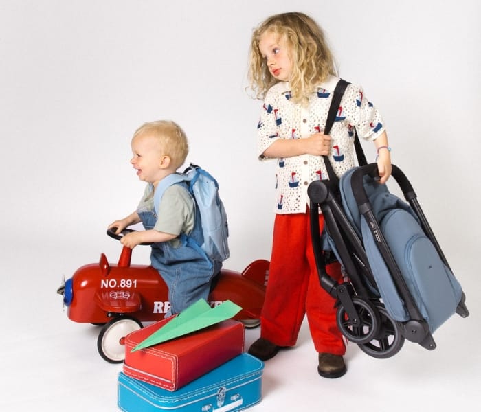 Child carrying a folded Joolz Aer stroller next to a toddler on a red toy airplane