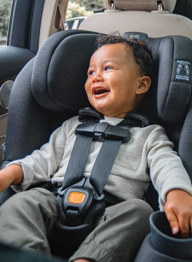 A baby with curly hair sits smiling in a gray car seat with black straps. "DUOGUARD HEAD & TORSO PROTECTION" text is visible.