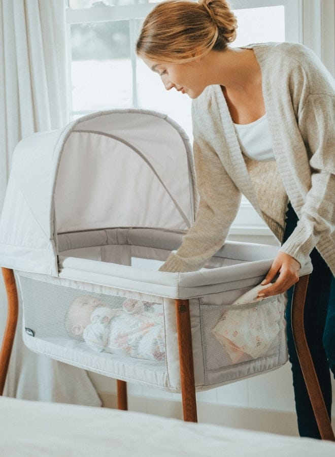 A woman tending to a baby in a light gray bassinet with mesh sides, set near a window with white curtains. She wears a cream-colored cardigan.