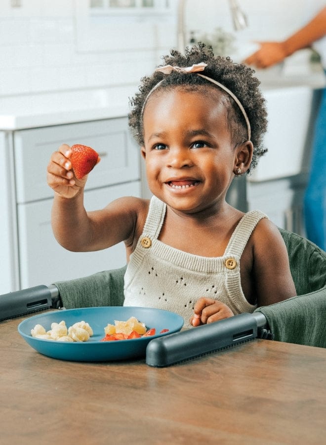 A toddler holds a strawberry, smiling in a highchair, with a plate of fruit in front of her on a wooden table.