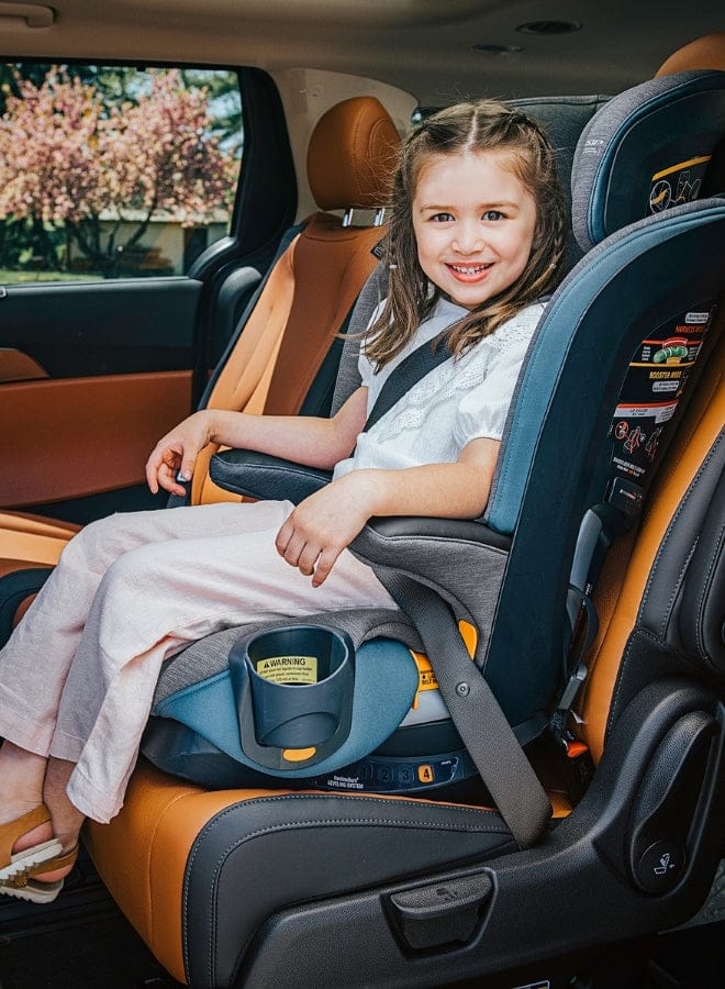 A smiling girl sits in a car seat, buckled in with a seatbelt. The car interior has brown and black leather seats, with a view to the outside.